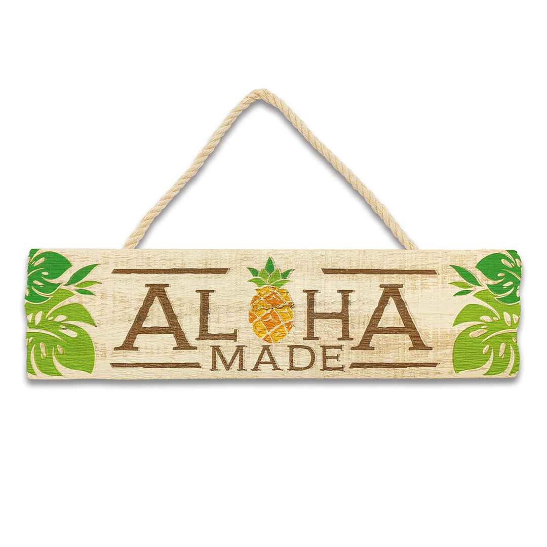 A wooden hanging sign with the text 'ALOHA MADE' carved out of a pineapple, featuring green leaves and a brown rope for hanging.