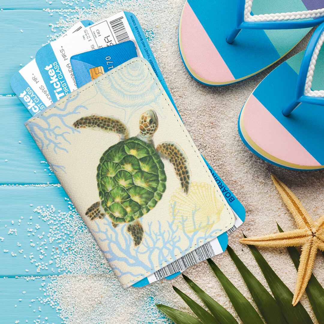 a lifestyle photo of a Hawaii sea turtle filled passport holder with beach sand and sandals