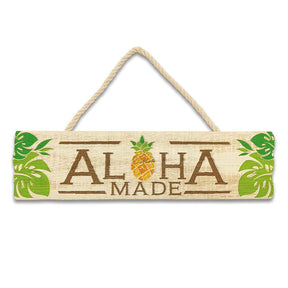 A wooden hanging sign with the text 'ALOHA MADE' carved out of a pineapple, featuring green leaves and a brown rope for hanging.