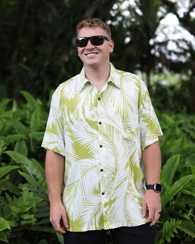 A man wearing a white short-sleeve button-up shirt with a green palm leaf pattern, black sunglasses, and a black smartwatch, standing in front of a lush green tropical background.