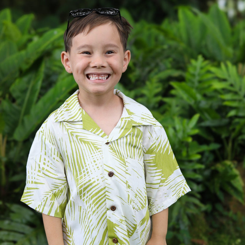 A young boy smiling outdoors in front of lush green foliage, wearing a white short-sleeve button-up shirt with a lime green palm leaf pattern and black sunglasses on his head.
